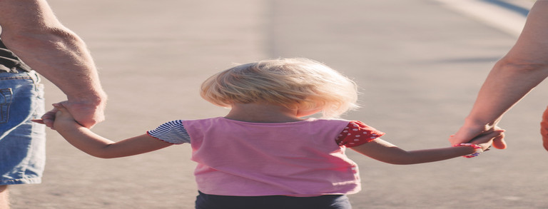 Rückansicht eines blonden Kindes, welches zwischen seinen Eltern läuft, ein Elternteil jeweils an einer Hand.