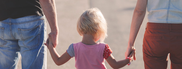 Rückansicht eines blonden Kindes, welches zwischen seinen Eltern läuft, ein Elternteil jeweils an einer Hand.