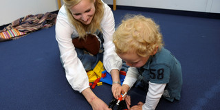 Spielsituation mit einer blonden Frau und einem Kind, die auf einem blauen Teppich sitzen und mit Spielzeug hantieren.