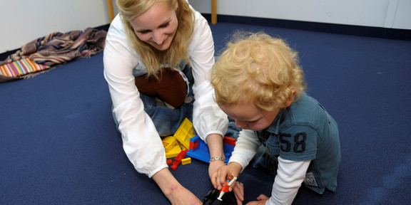 Spielsituation mit einer blonden Frau und einem Kind, die auf einem blauen Teppich sitzen und mit Spielzeug hantieren.