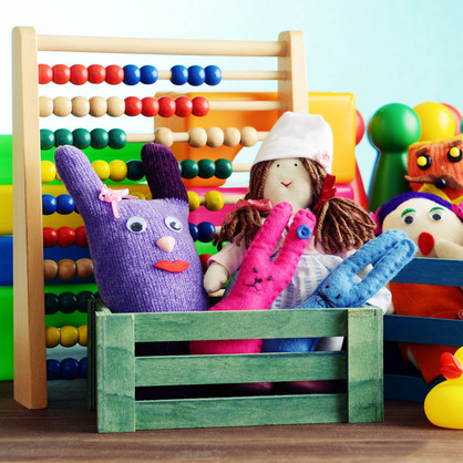 Eine Nahaufnahme verschiedener Spielzeuge in kleinen bunten Holzkisten. Man sieht Stoffpuppen, eine Gummiente, Spielfiguren und einen Abakus mit bunten Holzperlen.