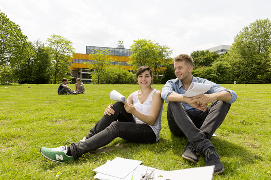 Zwei Studierende sitzen auf einer Wiese, im Hintergrund die Mensa